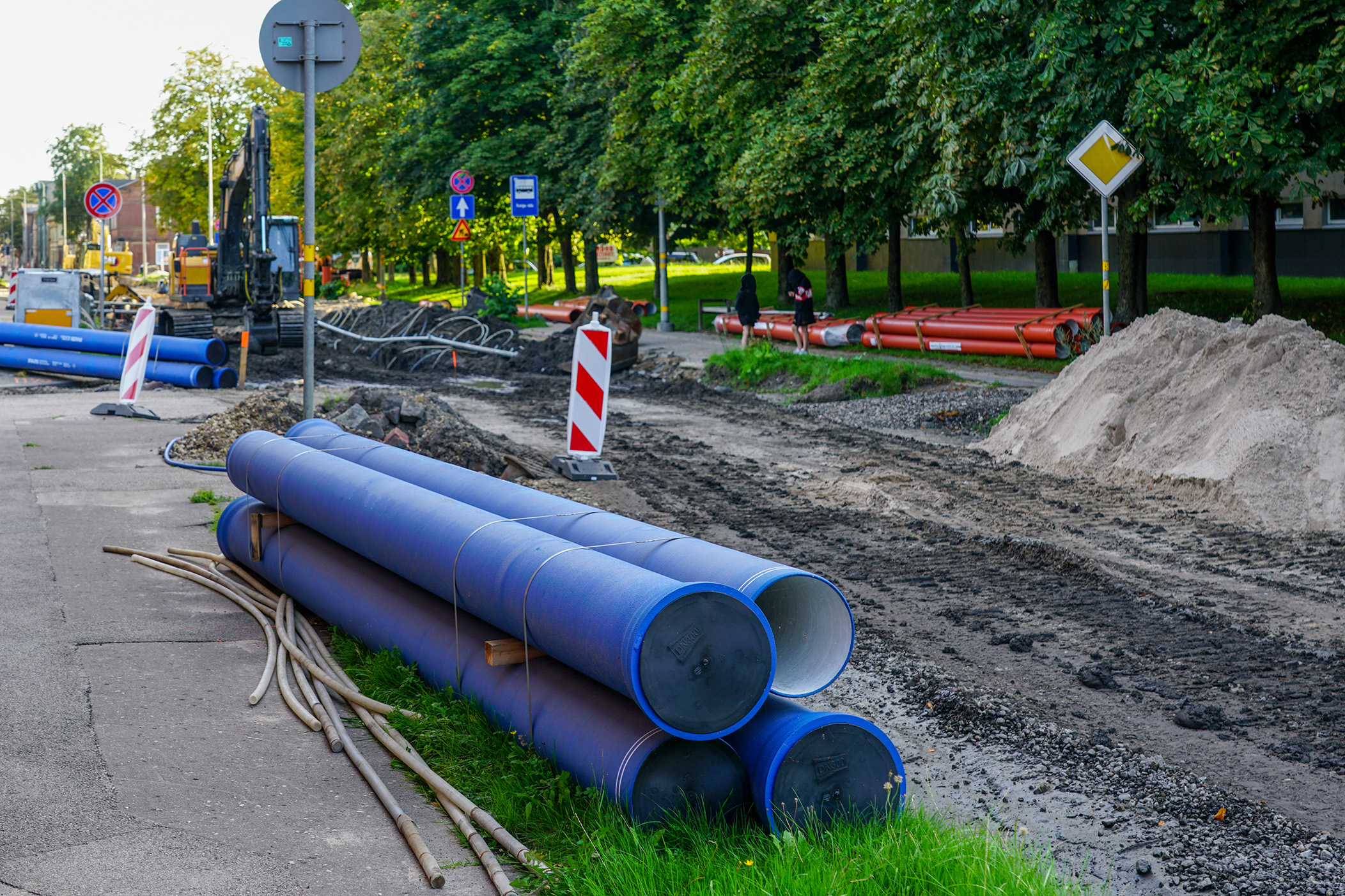 Browne Group - Blue pipes and construction materials by a dug-up road with traffic barriers and green trees nearby.