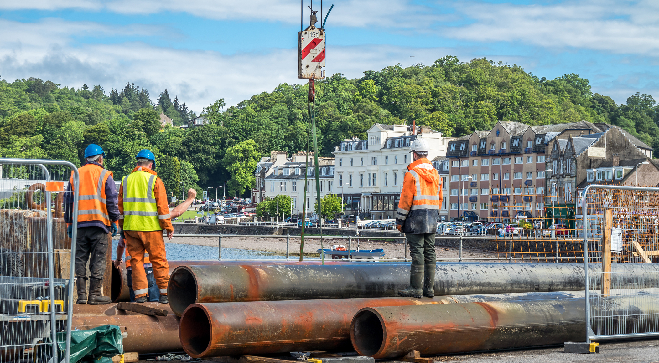 Browne Group - Construction workers in high-visibility gear stand near large metal pipes by a waterfront in a town.