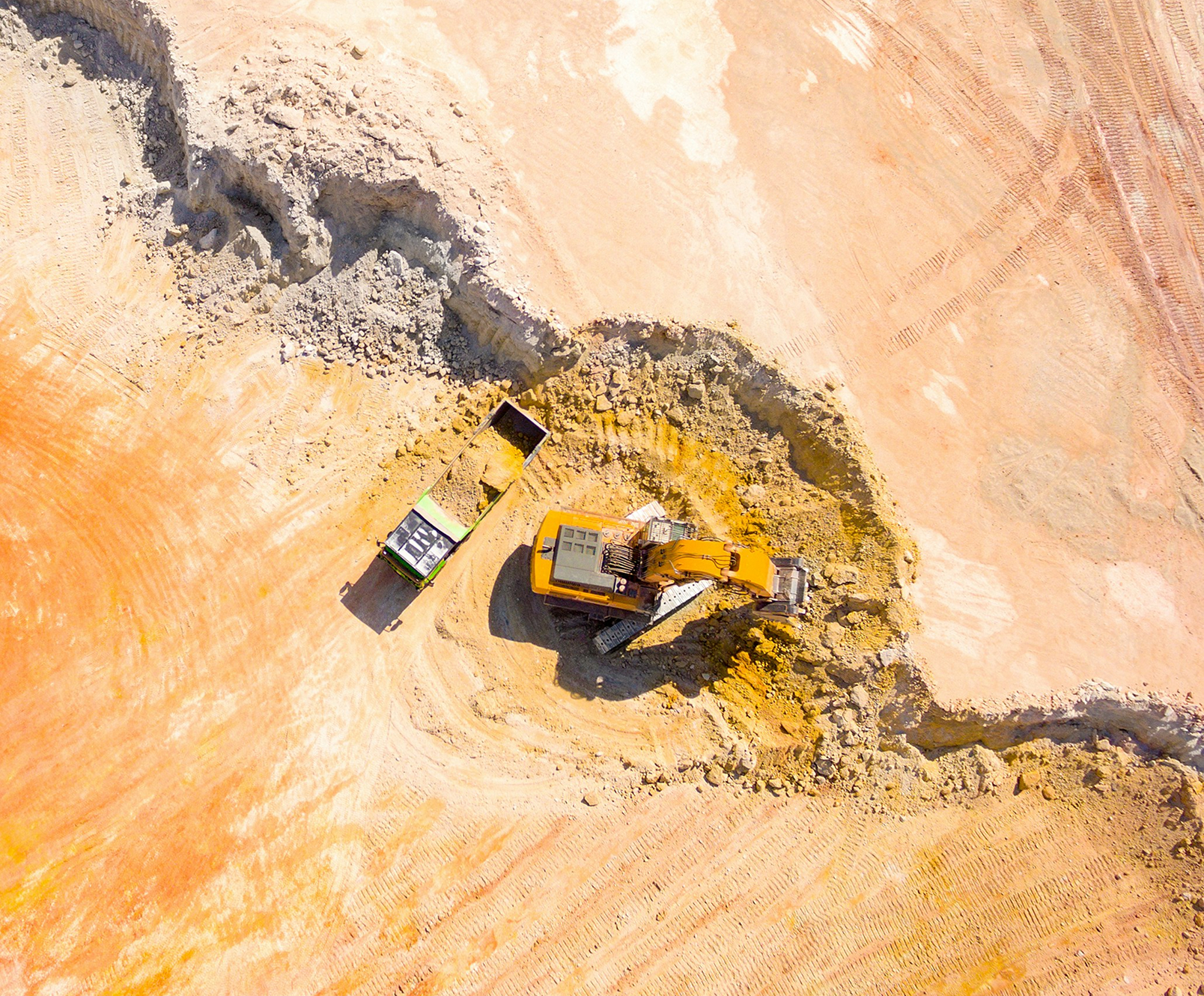 Browne Group - Aerial view of a yellow excavator loading dirt into a truck on a construction site with reddish soil.