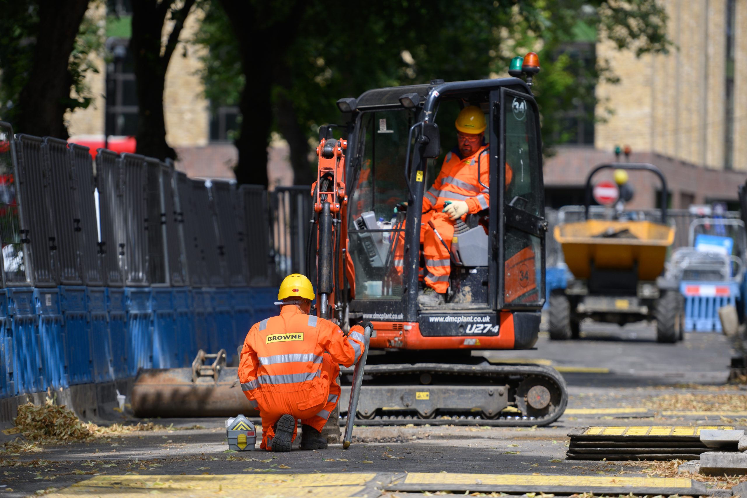 Browne Group - Two construction workers in orange safety gear work on a city street with a mini excavator and barriers.