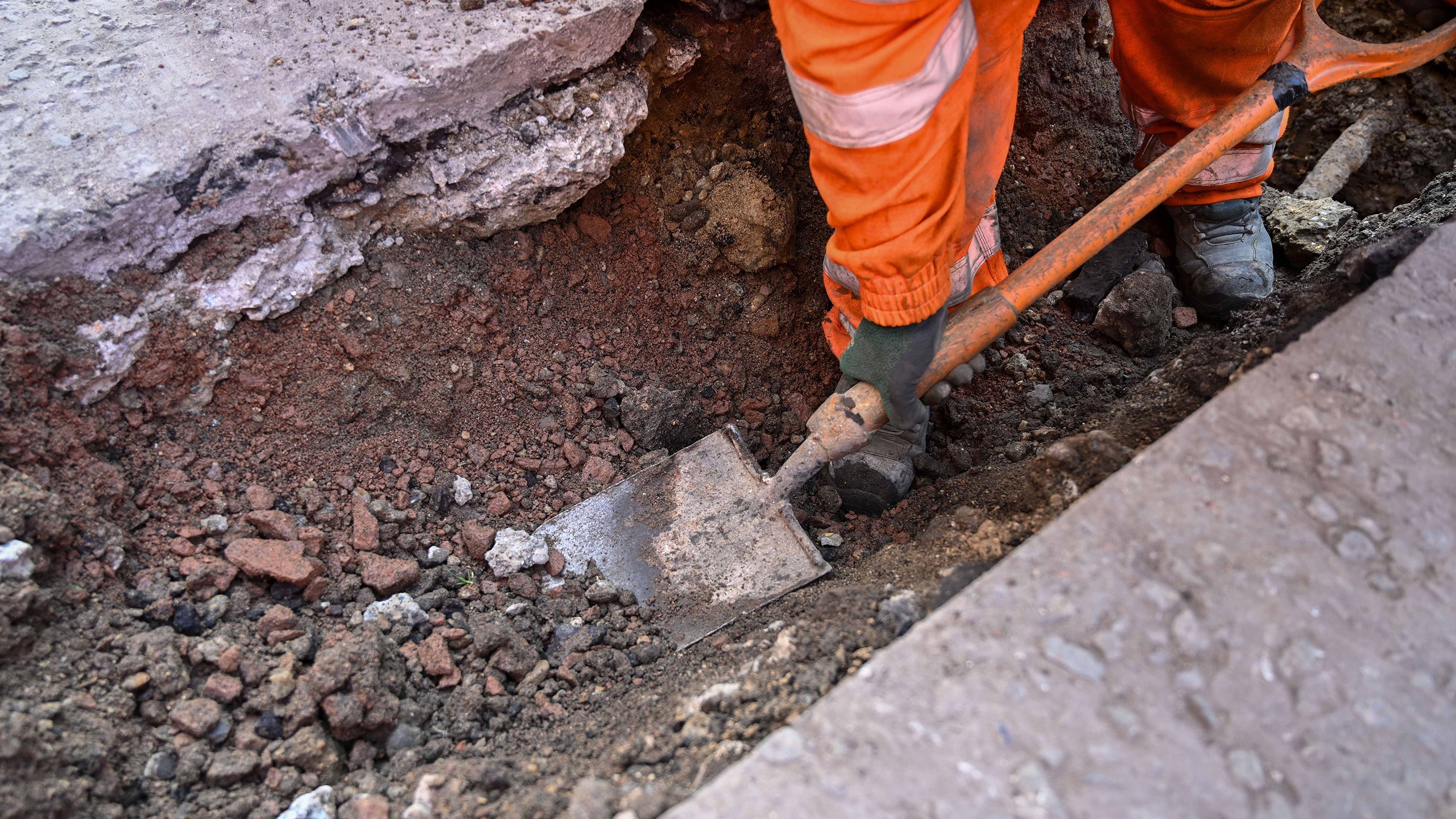 Browne Group - Worker in orange pants digging with a shovel at a construction site with loose soil and rubble.