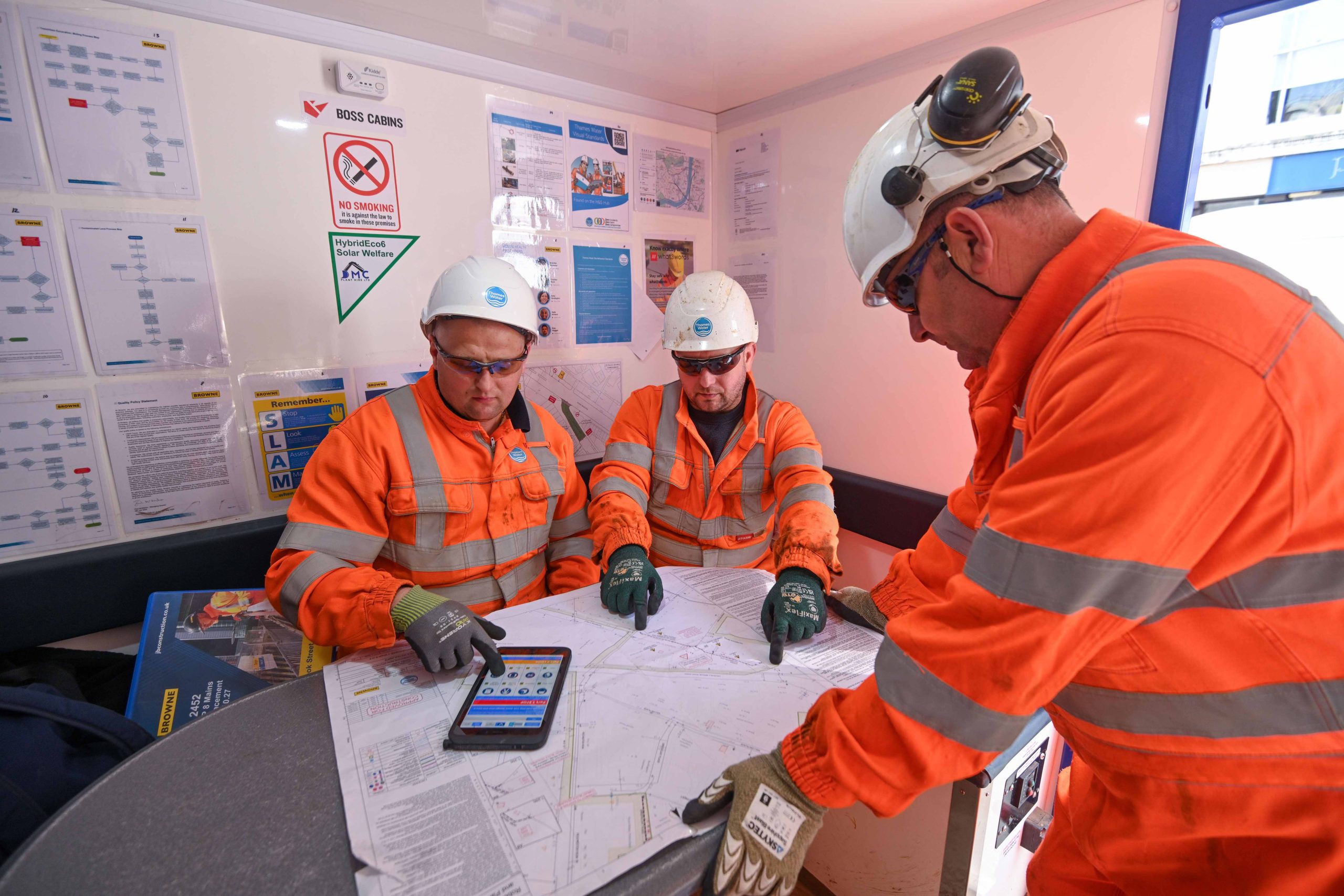 Browne Group - Three construction workers in orange safety gear and helmets review plans and a tablet together at a worksite table.