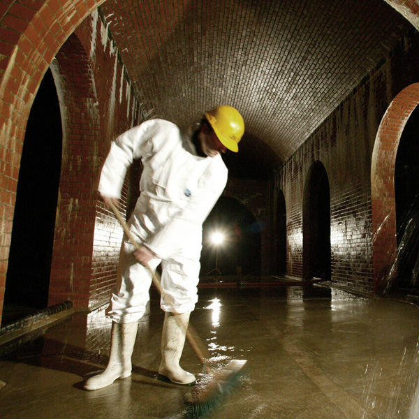 Browne Group - Person in white protective suit and yellow helmet cleaning a wet floor in an arched brick tunnel.