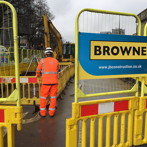 Browne Group - Construction worker in orange gear stands by yellow barriers at a roadwork site with machinery and signage.