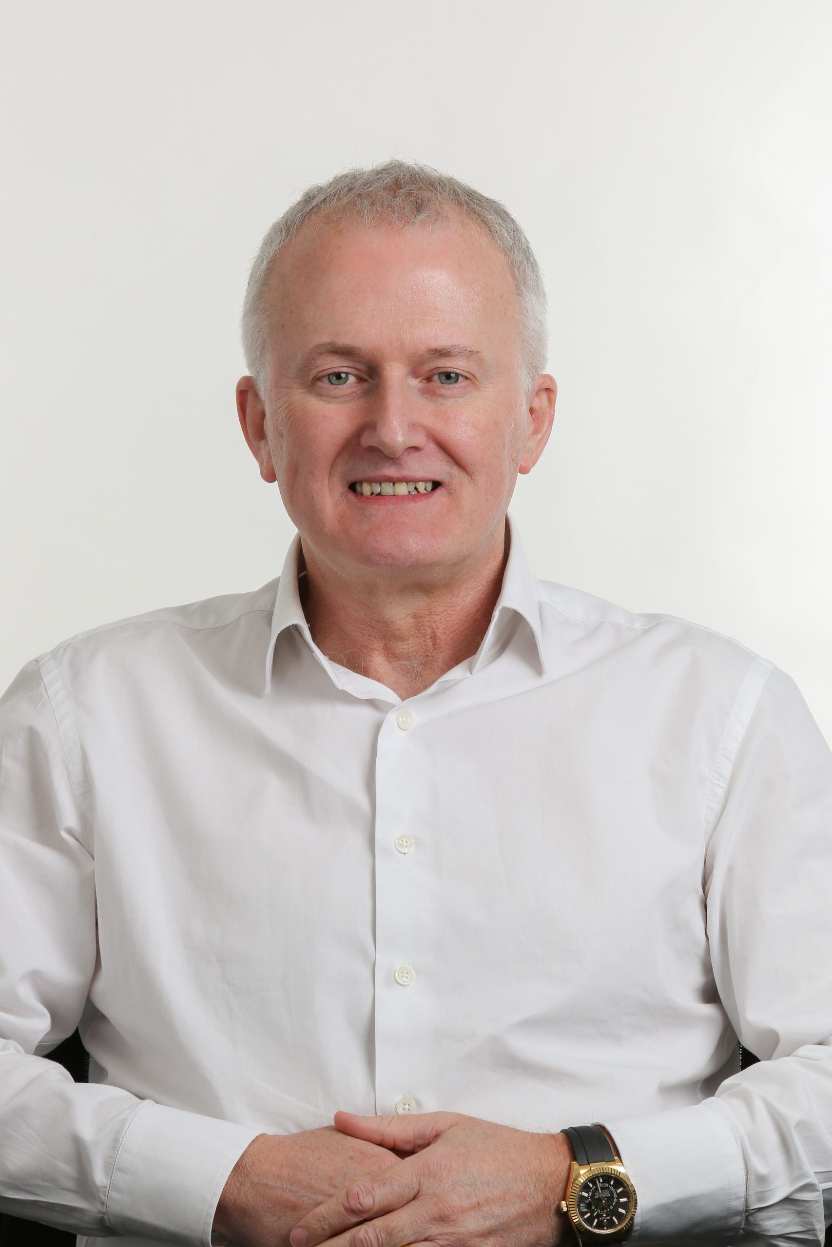 Browne Group - Smiling man in a white shirt sits with hands folded, against a plain light background.