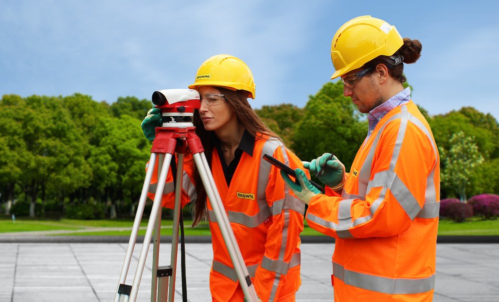 Browne Group - Two surveyors in orange safety gear use surveying equipment outdoors near trees on a clear day.