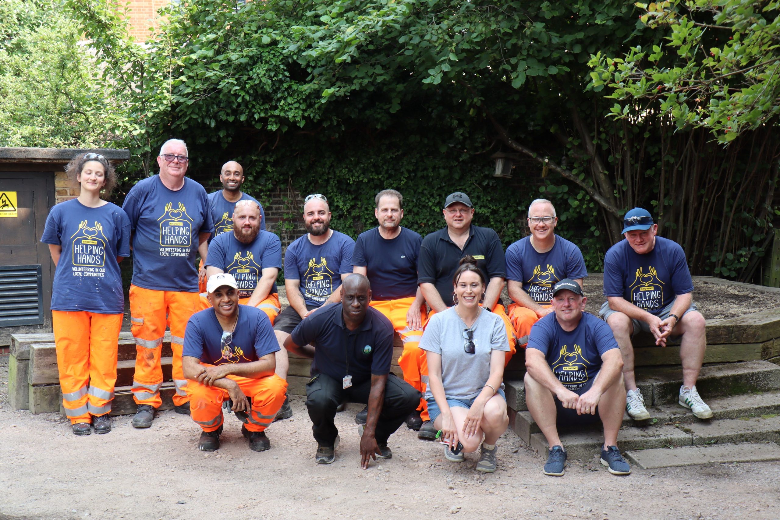 Browne Group - A group of twelve people wearing work clothes and smiling, posing outdoors in front of greenery and steps.