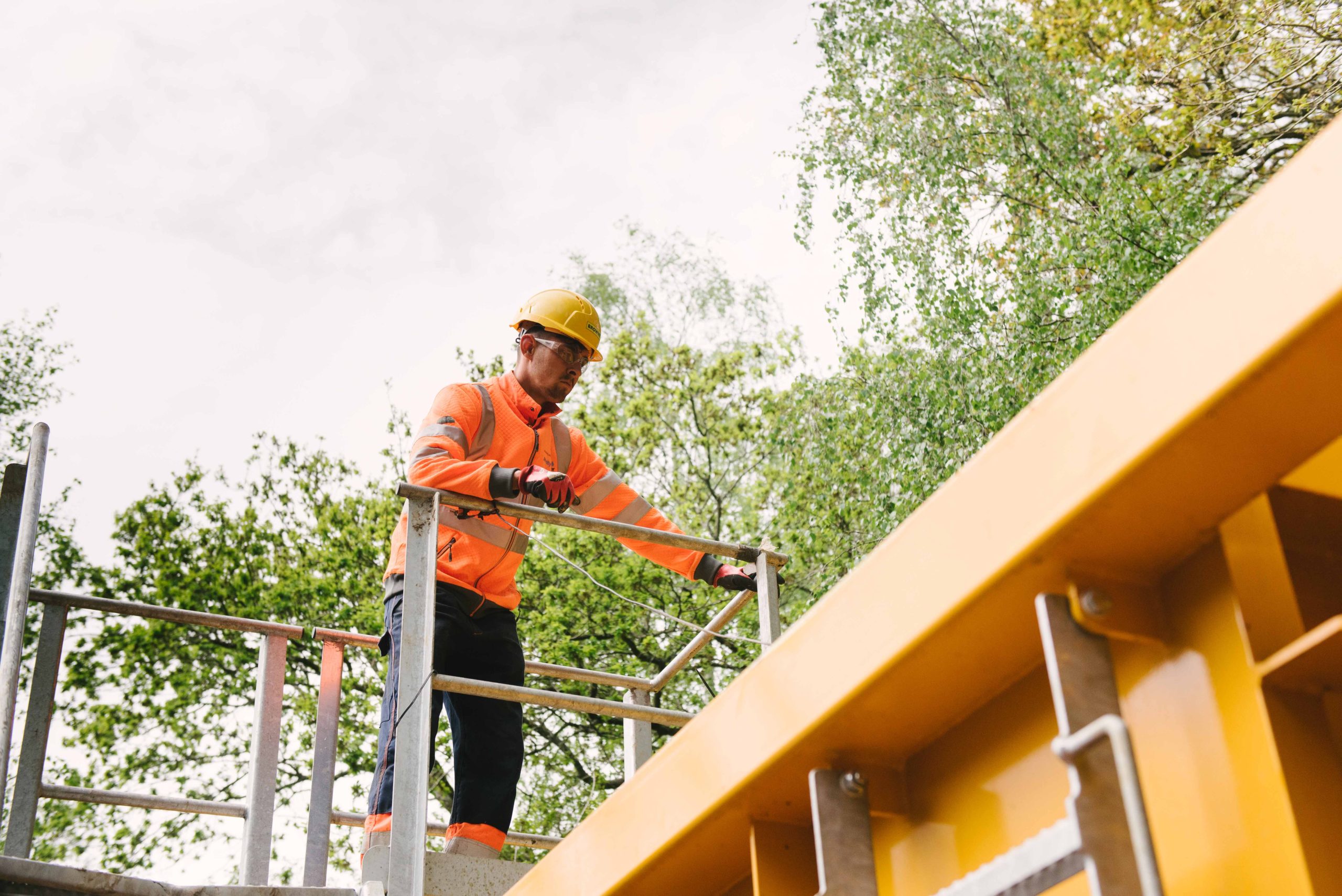 Browne Group - Construction worker in a hard hat and orange vest standing on scaffolding outdoors near trees.
