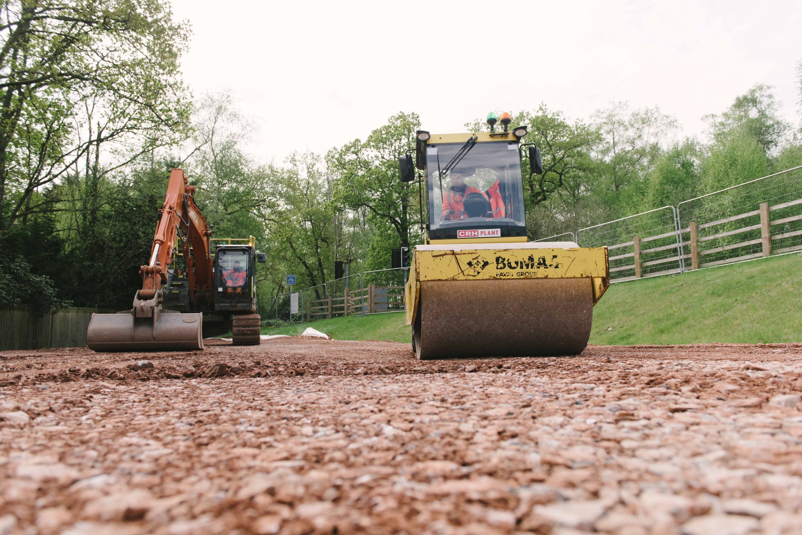 Browne Group - A yellow steamroller flattens a dirt road near trees, with an excavator working behind it.