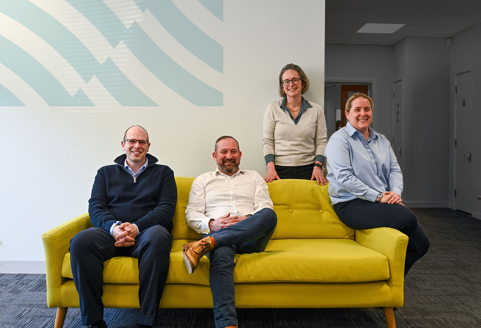 Browne Group - Four people, two seated on a yellow sofa and two standing behind it, smiling in a modern office space.