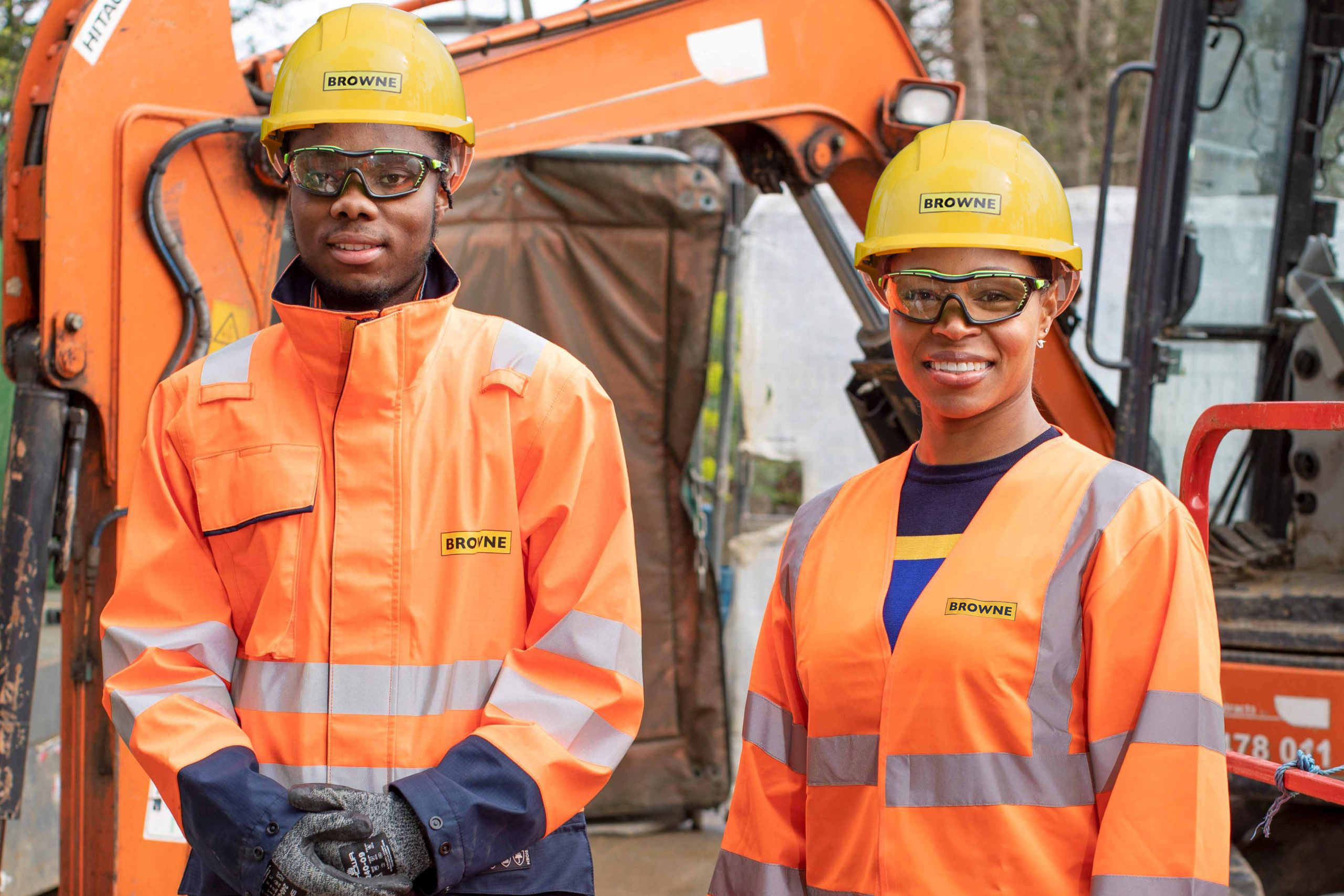 Browne Group - Two construction workers in orange safety gear and helmets stand near an excavator, smiling at the camera.