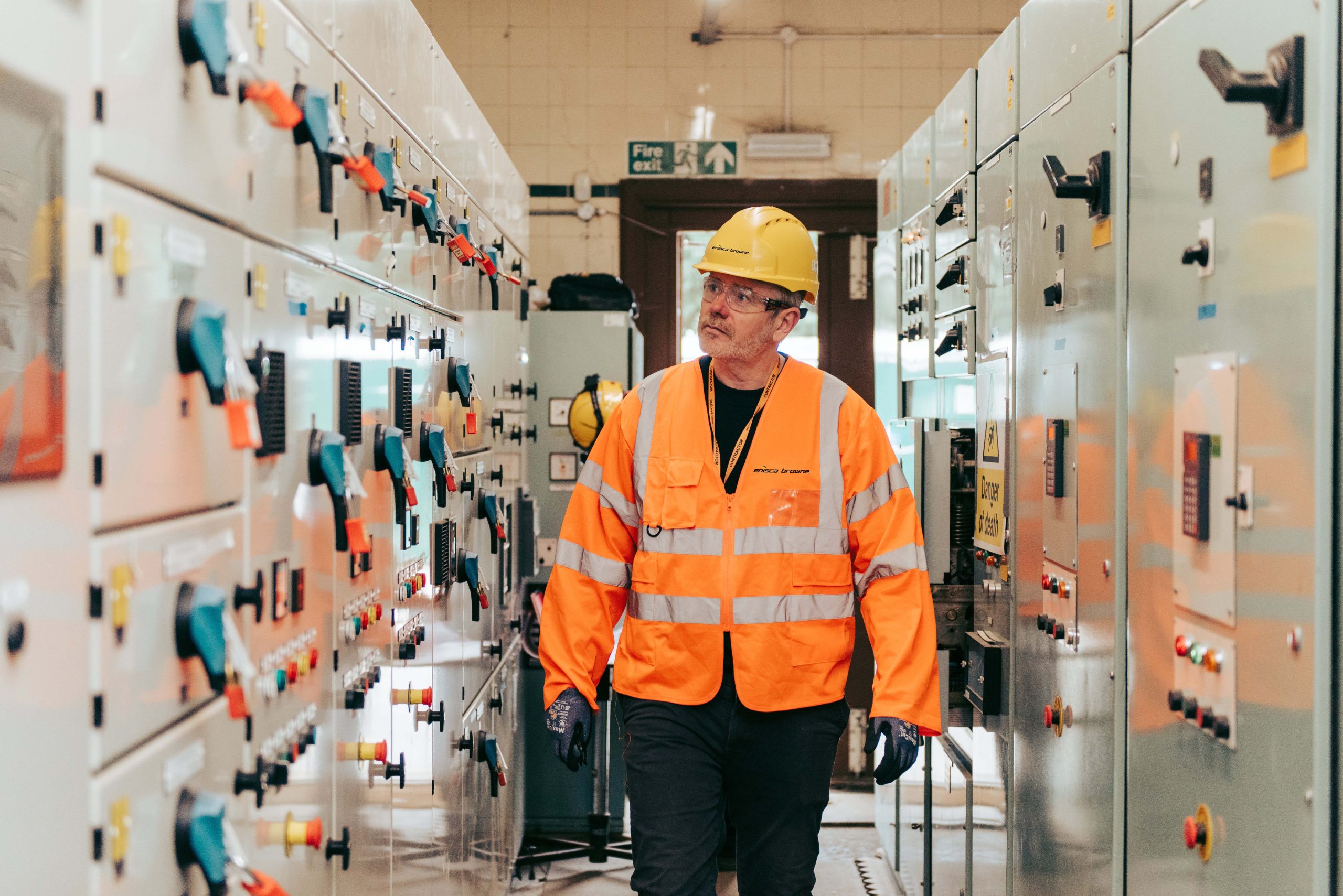 Browne Group - A worker in a hard hat and orange safety vest inspects control panels in an industrial facility.