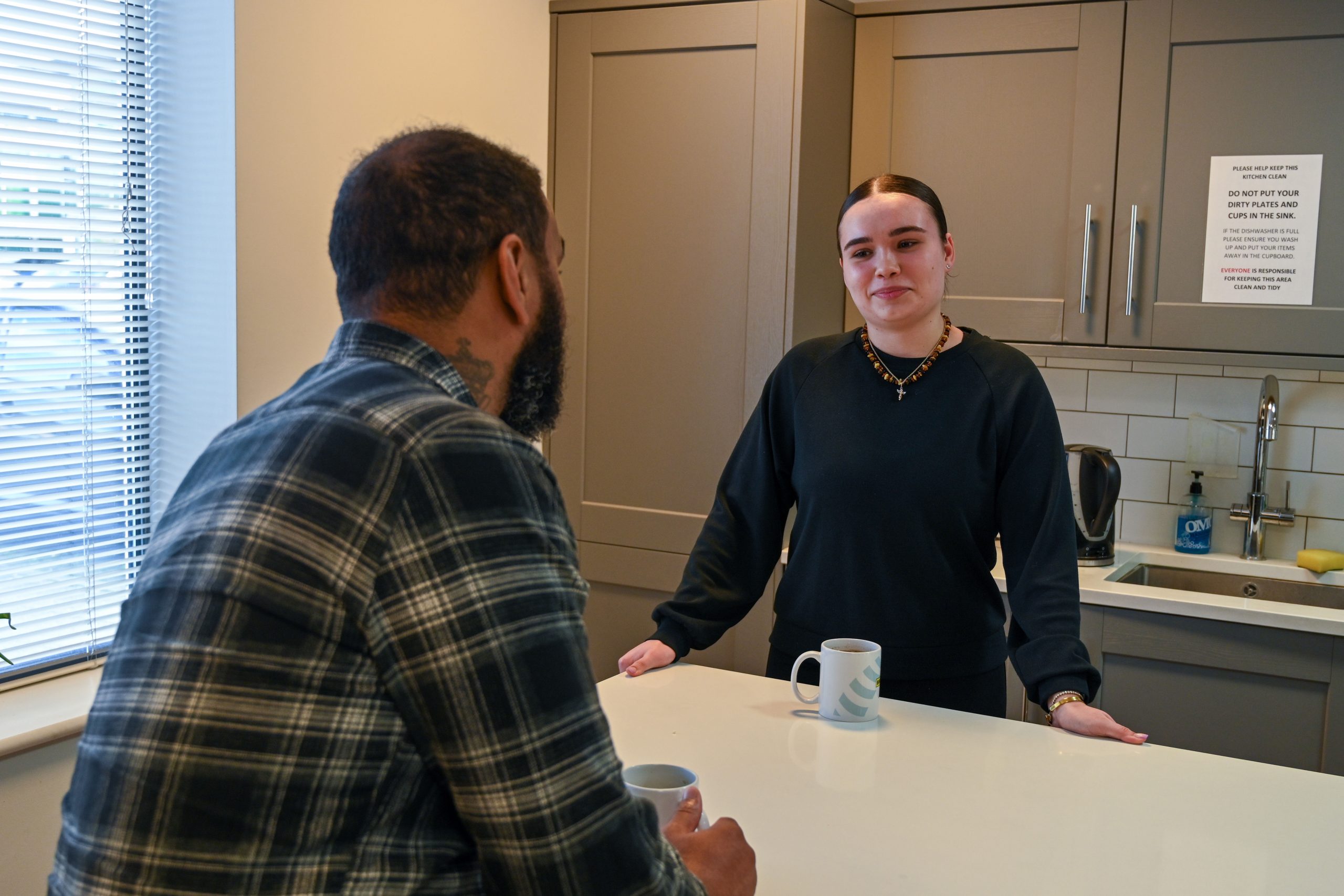 Browne Group - Two people talking in a kitchen, each holding a mug, standing by a counter near a window and cabinets.