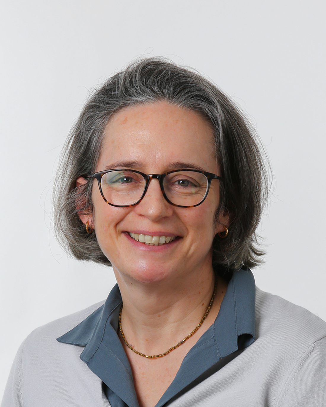 Browne Group - Woman with short gray hair and glasses, smiling, wearing a gray sweater over a blue collared shirt.