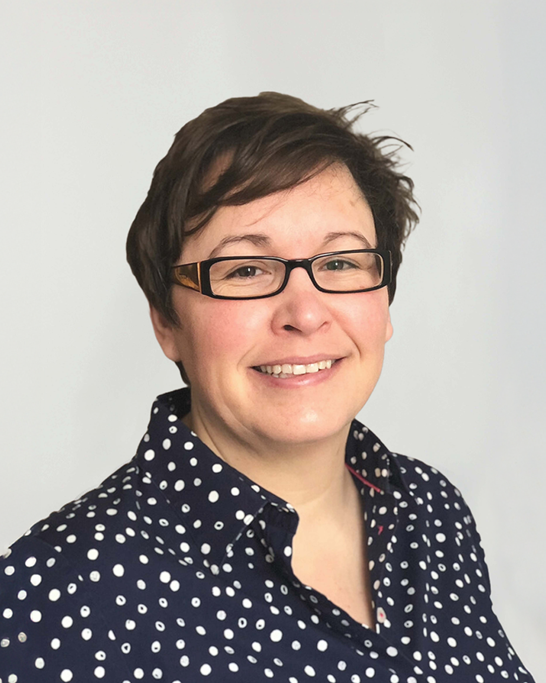 Browne Group - Smiling person with short brown hair and glasses, wearing a navy blue polka dot shirt, against a light background.