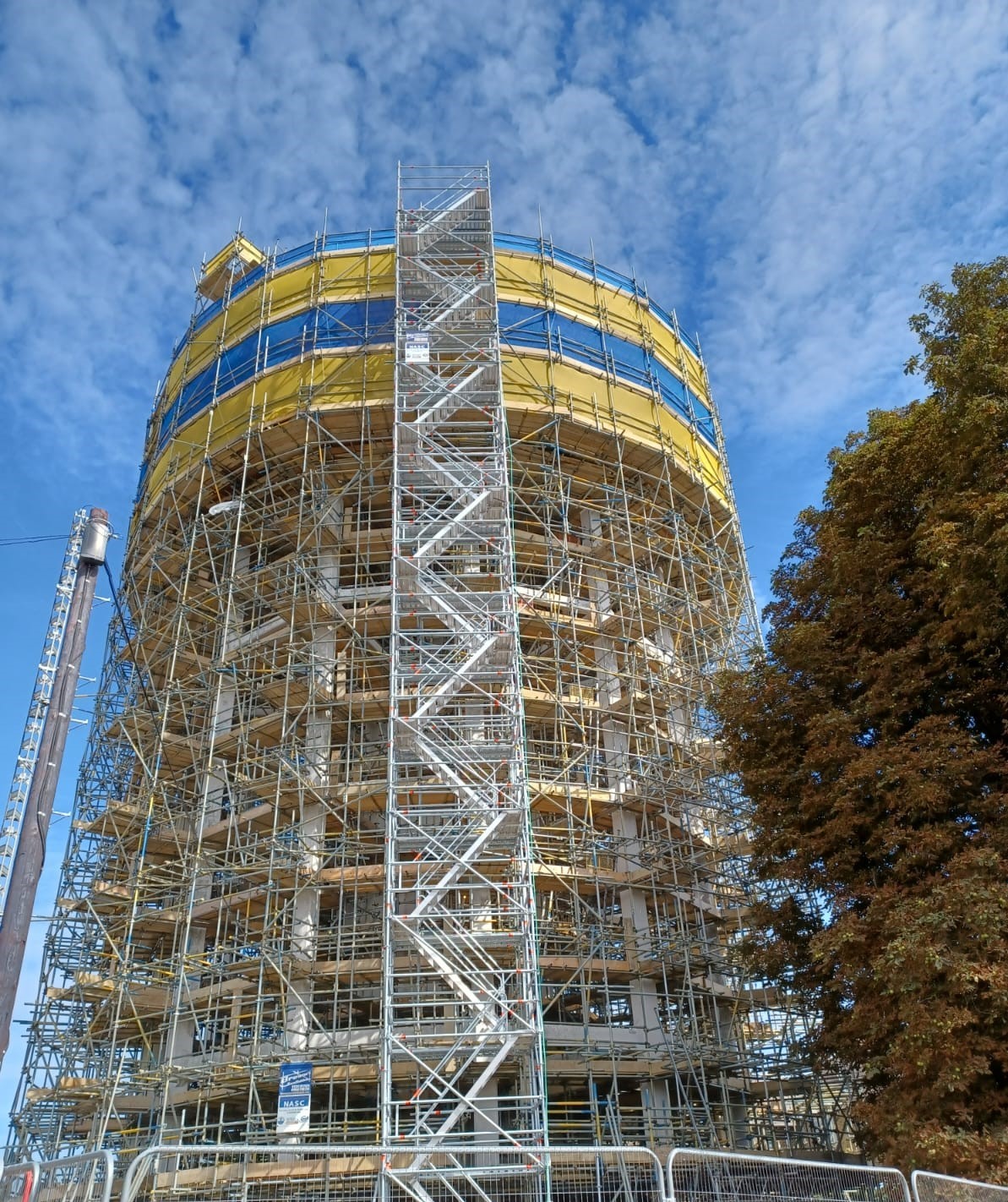 Browne Group - A round building covered in scaffolding with yellow covers under a blue sky, next to a tree.