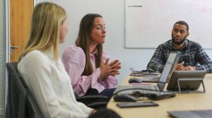 Browne Group - Three people sit at a conference table having a meeting, with laptops and a whiteboard in the background.