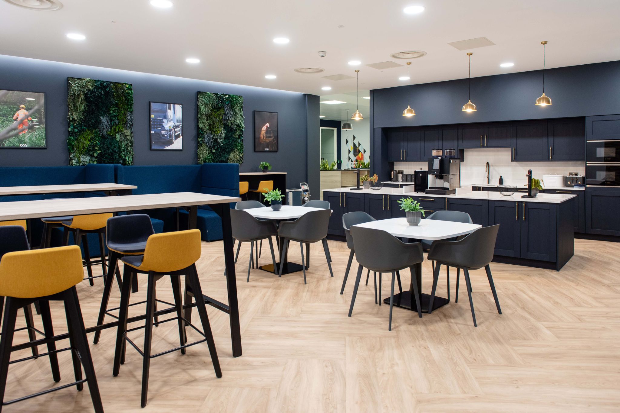 Browne Group - Modern office kitchen with tables, chairs, plants, dark cabinets, wall art, and pendant lights.