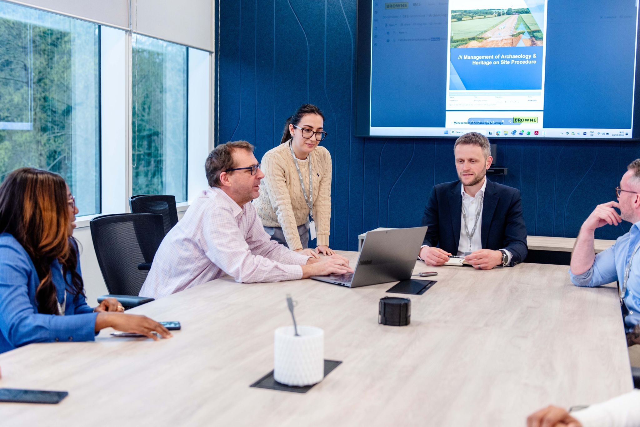 Browne Group - Five people have a meeting around a table with a laptop, with a presentation on a screen in the background.
