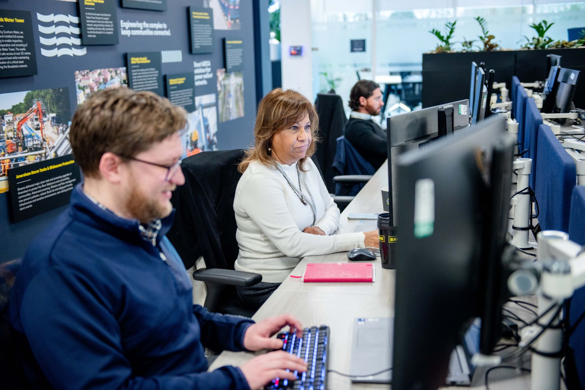 Browne Group - Three people work at computers in a modern office with informational posters on the wall and plants in the background.