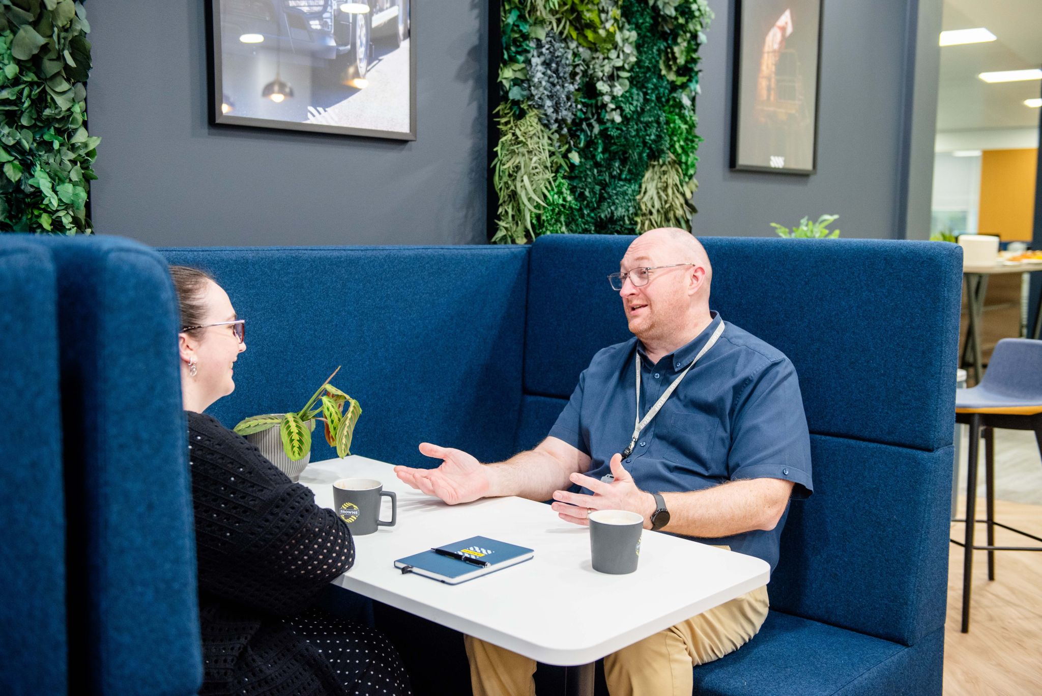 Browne Group - Two people sit in a modern office booth, talking over coffee, notebooks, and a small plant on the table.