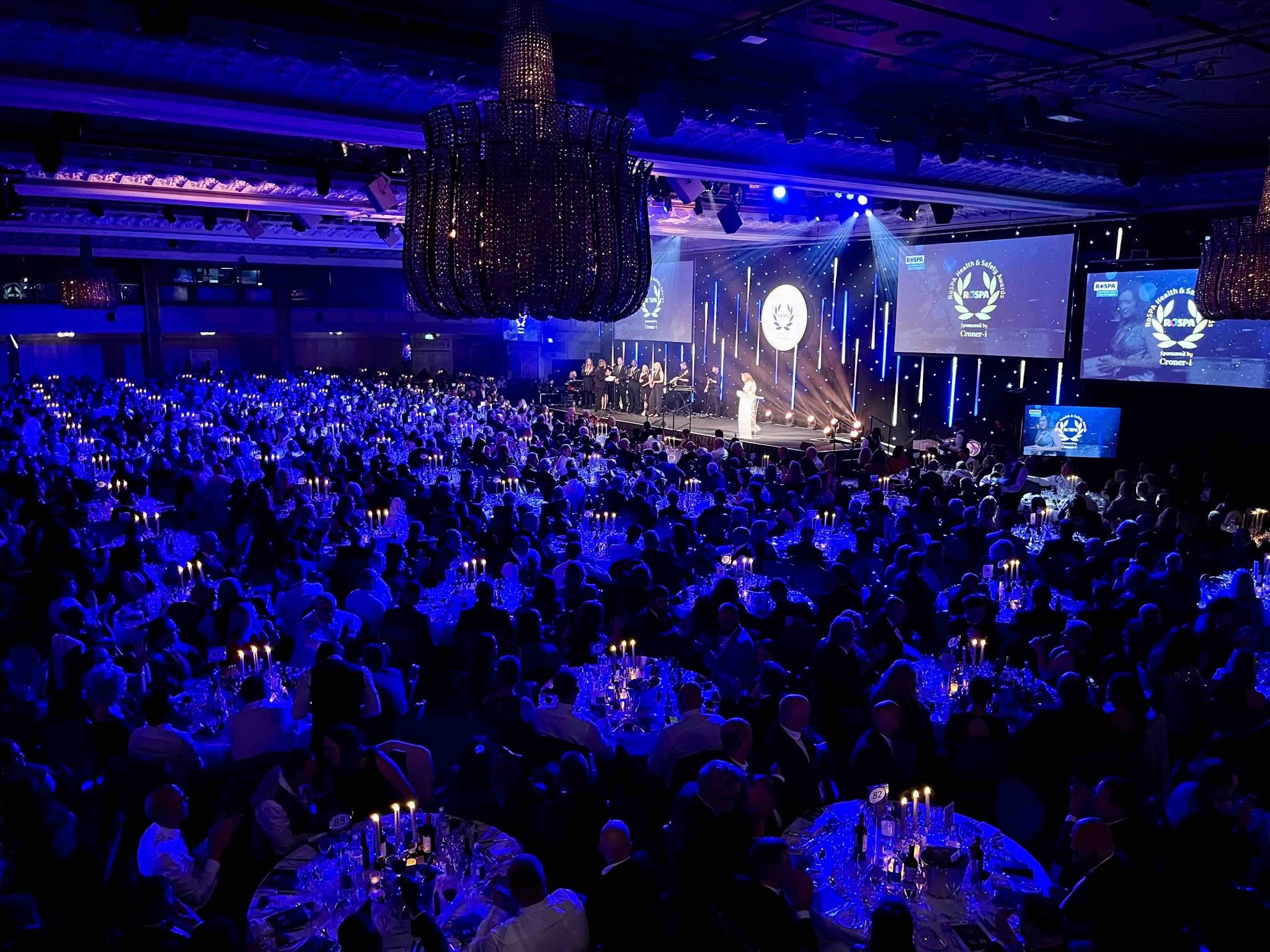 Browne Group - A large audience seated at round tables in a ballroom watches a person speaking on a lit stage with blue lighting.