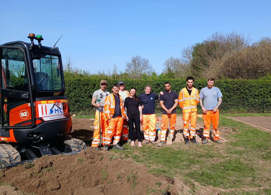 Browne Group - Seven people in orange work pants stand outdoors by an excavator, smiling, with green trees and blue sky behind them.