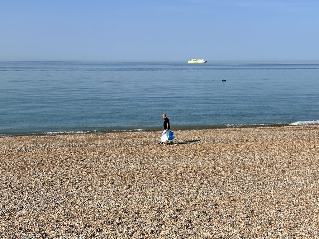 Browne Group - A person collects trash on a rocky beach during the Thames Water Raft Race, with calm water and a yellow boat visible.