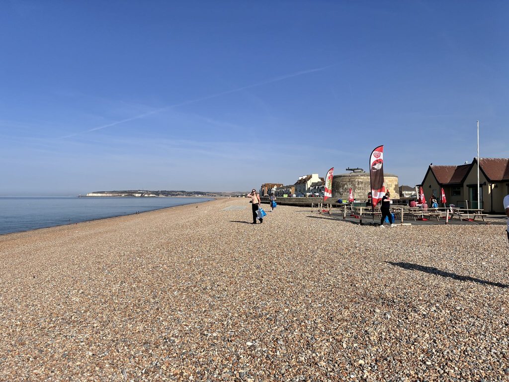 Browne Group - A pebbly beach with a few people, flags, huts, and calm sea hosts the lively Thames Water Raft Race under clear skies.